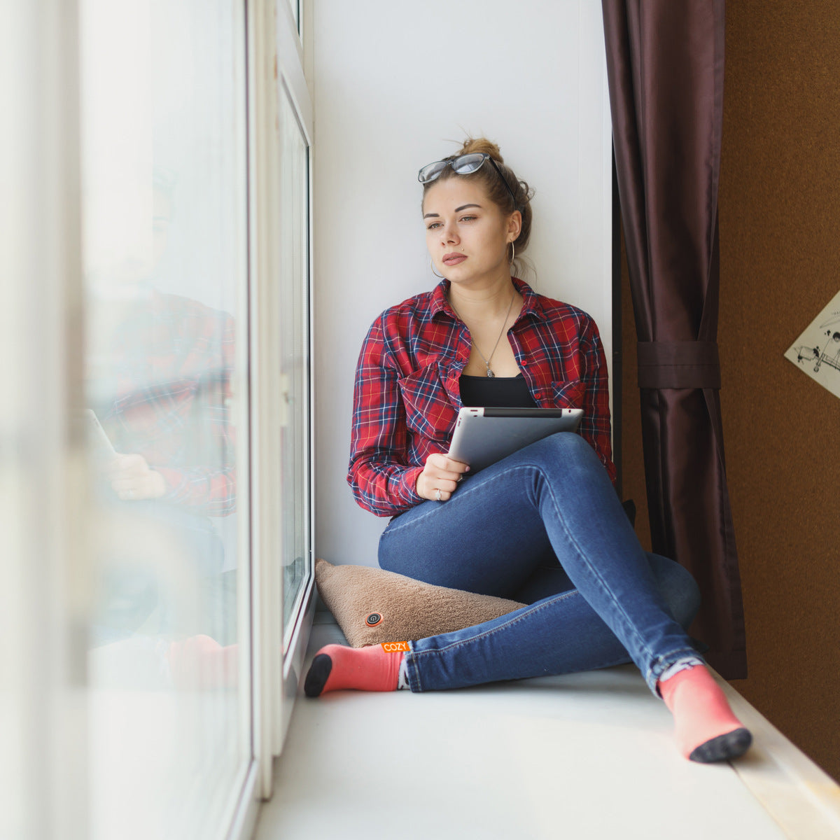A woman in a red plaid shirt and jeans sits on a windowsill, holding a tablet, relaxing with the COZY Heated Cushion Cordless - UNA (45cm x 45cm) by Cozy Cushion EU, enjoying cordless warmth as sunlight streams in.