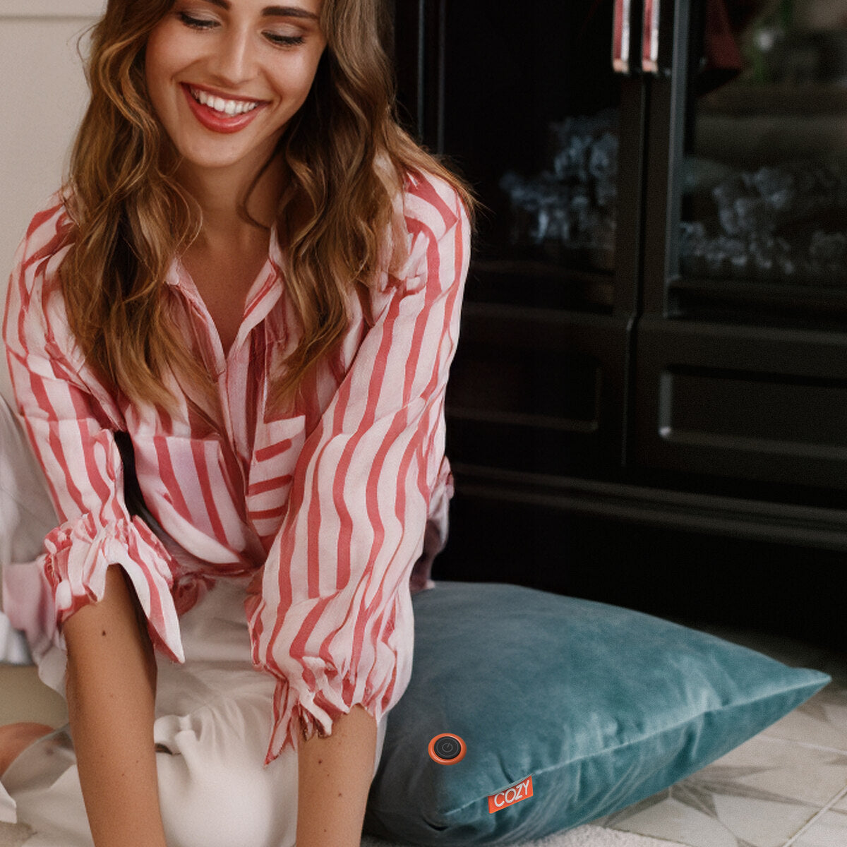 A woman with wavy hair, wearing a pink and white striped blouse, sits smiling on the floor beside a teal COZY Heated Cushion Cordless - UNA Velvet (45cm x 45cm) by Cozy Cushion EU. The cushion has an orange “COZY” tag and runs on a rechargeable battery. A black fireplace is behind her.