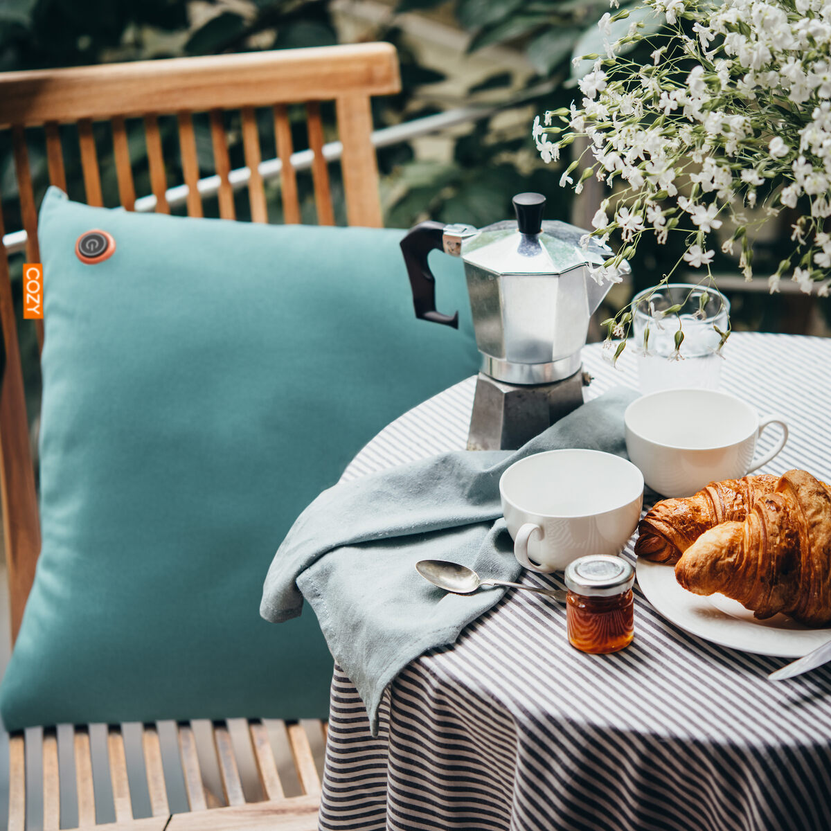 A cozy outdoor breakfast scene with a striped tablecloth, two white cups, a glass, croissants, a moka pot, and the COZY Heated Cushion Cordless - UNA Velvet (45cm x 45cm) by Cozy Cushion EU on a wooden chair beside a vase of white flowers.