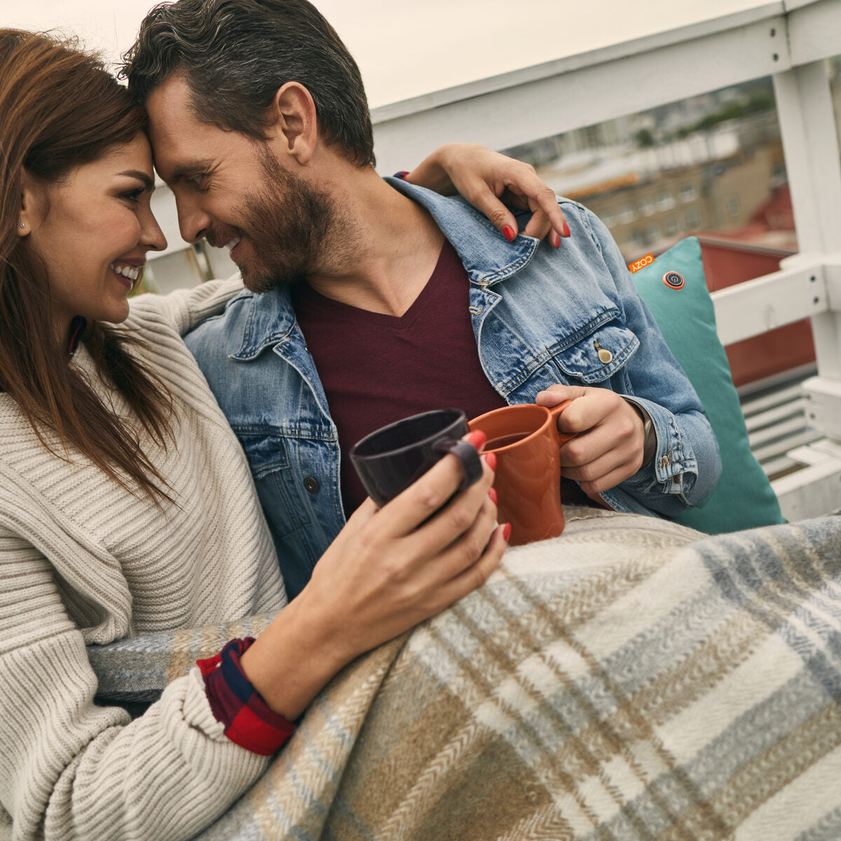 A smiling couple cuddles under a plaid blanket on a rooftop, enjoying coffee and the warmth of the COZY Heated Cushion Cordless - UNA Velvet (45cm x 45cm) by Cozy Cushion EU, powered by cordless infrared technology.