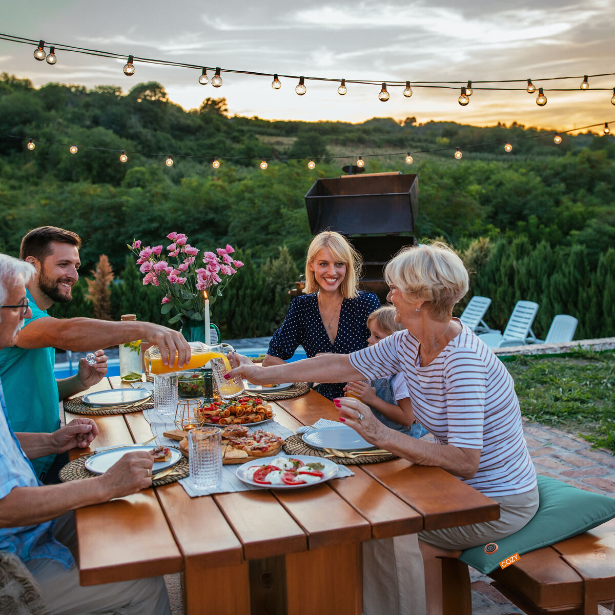A group enjoys a meal at sunset on Cozy Cushion EU’s COZY Heated Cushion Cordless - UNA Velvet (45cm x 45cm), sitting around a wooden picnic table outdoors under string lights, surrounded by greenery and a scenic view.