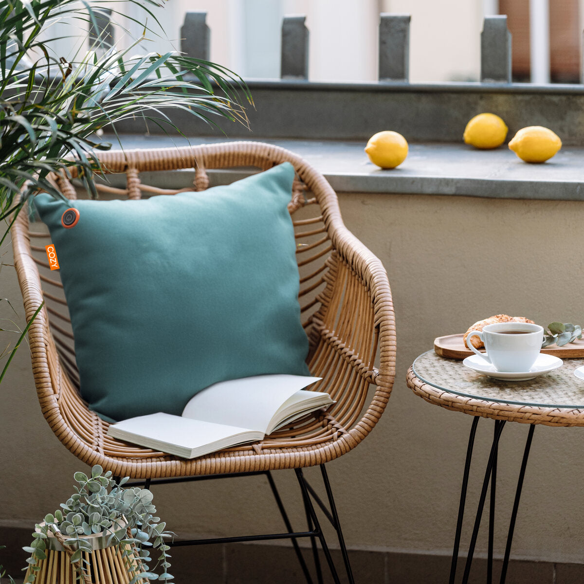 A rattan chair with the COZY Heated Cushion Cordless - UNA Velvet (45cm x 45cm) by Cozy Cushion EU and an open book sits on a balcony beside a round table with coffee, a plant, and cookies. Three lemons are on the ledge behind the chair.