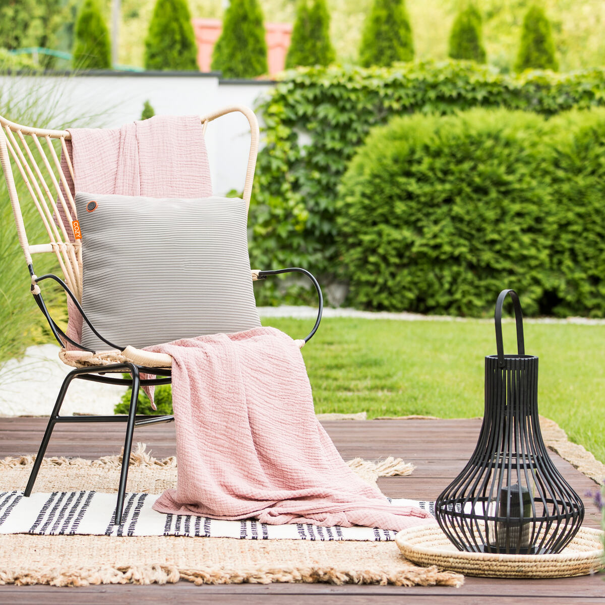 A cozy outdoor seating area with a chair, draped in a pink blanket and gray pillow, features the COZY Heated Cushion Cordless - UNA Knitted (45cm x 45cm) by Cozy Cushion EU, plus a black lantern on a woven mat and patterned rug on the deck amid lush greenery.