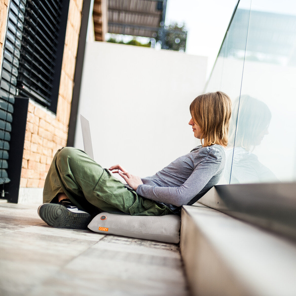 A person sits against a glass wall, working on a laptop while using the COZY Heated Cushion Cordless - UNA Knitted (45cm x 45cm) from Cozy Cushion EU. They wear green pants and a gray long-sleeve shirt as sunlight streams onto the tiled floor.