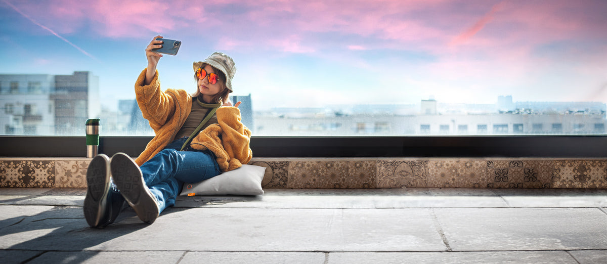 A person in a yellow jacket and sunglasses sits on the COZY Heated Cushion Cordless - UNA Knitted (45cm x 45cm) by Cozy Cushion EU near a large window, taking a selfie with a smartphone; a travel mug and pink-blue sky are nearby.