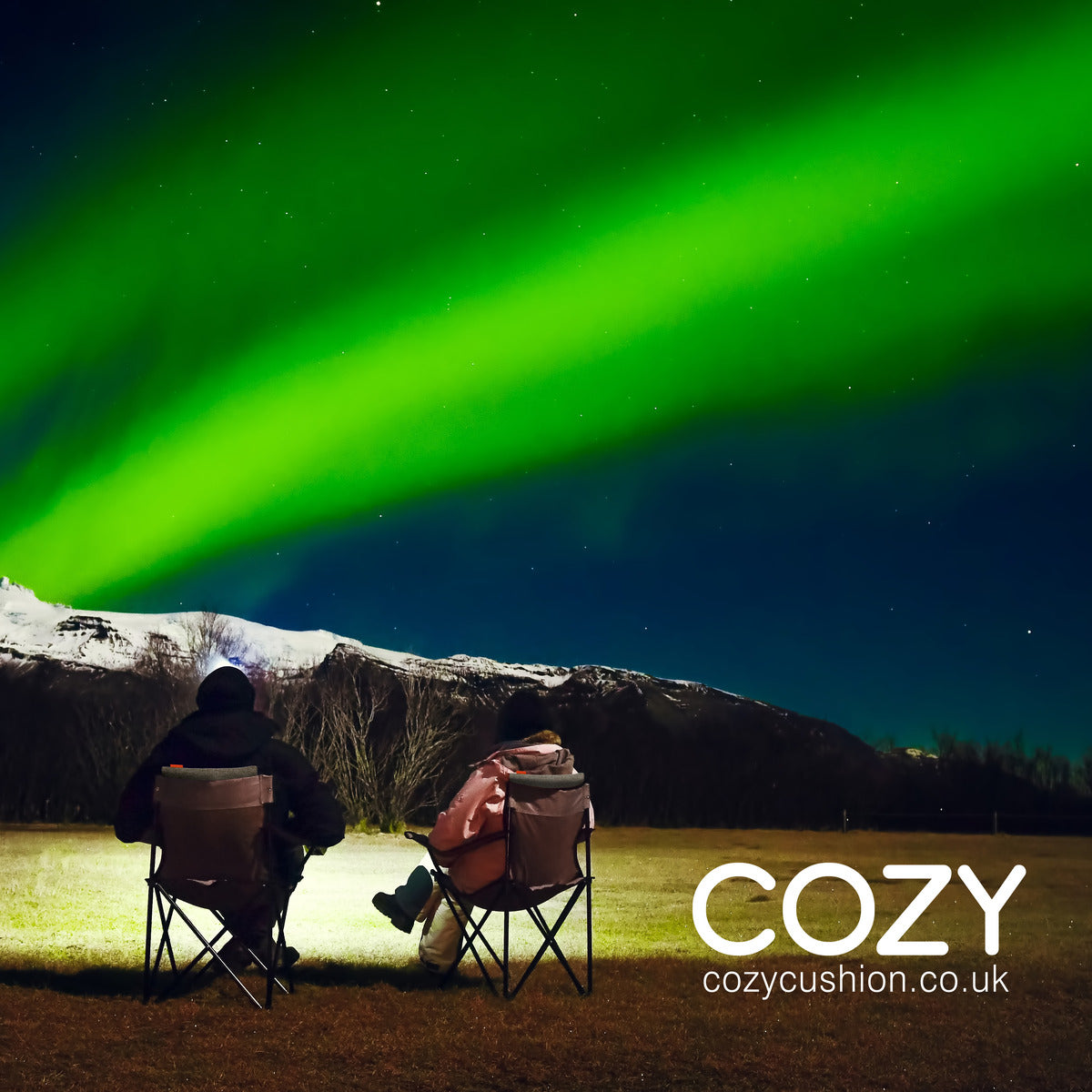 Two people in winter clothing sit on chairs under green northern lights, staying warm with the COZY Heated Cushion Cordless - UNA Knitted by Cozy Cushion EU. Text reads COZY and cozycushion.co.uk appears in the bottom right corner.