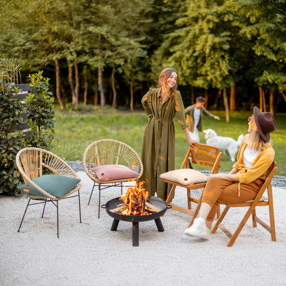 Two women relax on stylish chairs with Cozy Cushion EU’s COZY Heated Cushion Cordless - UNA Faux Fur (45cm x 45cm) by a fire pit in a garden. Nearby, a child and white dog play on the grass, surrounded by lush bushes and trees.