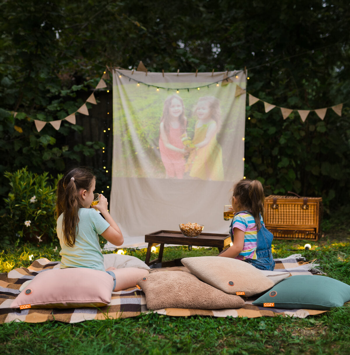 Two young girls relax outdoors on pillows and a COZY Heated Cushion Cordless - UNA Velvet (45cm x 45cm) by Cozy Cushion EU, watching a movie under string lights with snacks nearby. Trees and greenery provide a cozy atmosphere.