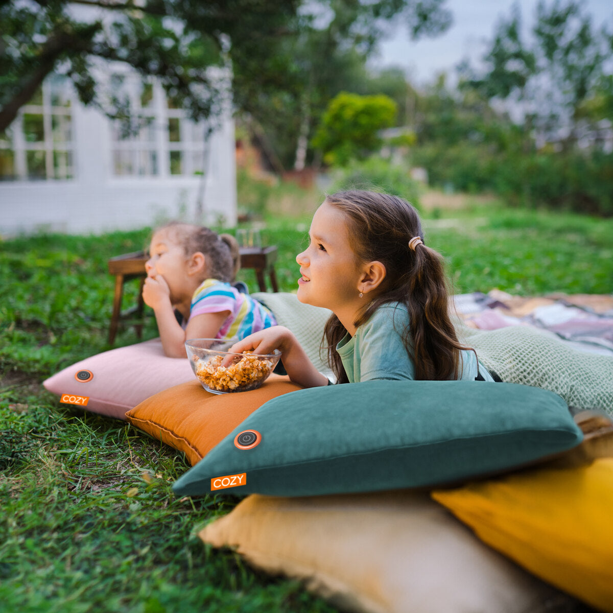 Two young girls relax on the grass, enjoying popcorn while lounging on COZY Heated Cushion Cordless – UNA Velvet (45cm x 45cm) from Cozy Cushion EU. The rechargeable cushions keep them warm outdoors, with greenery and a house in the background.