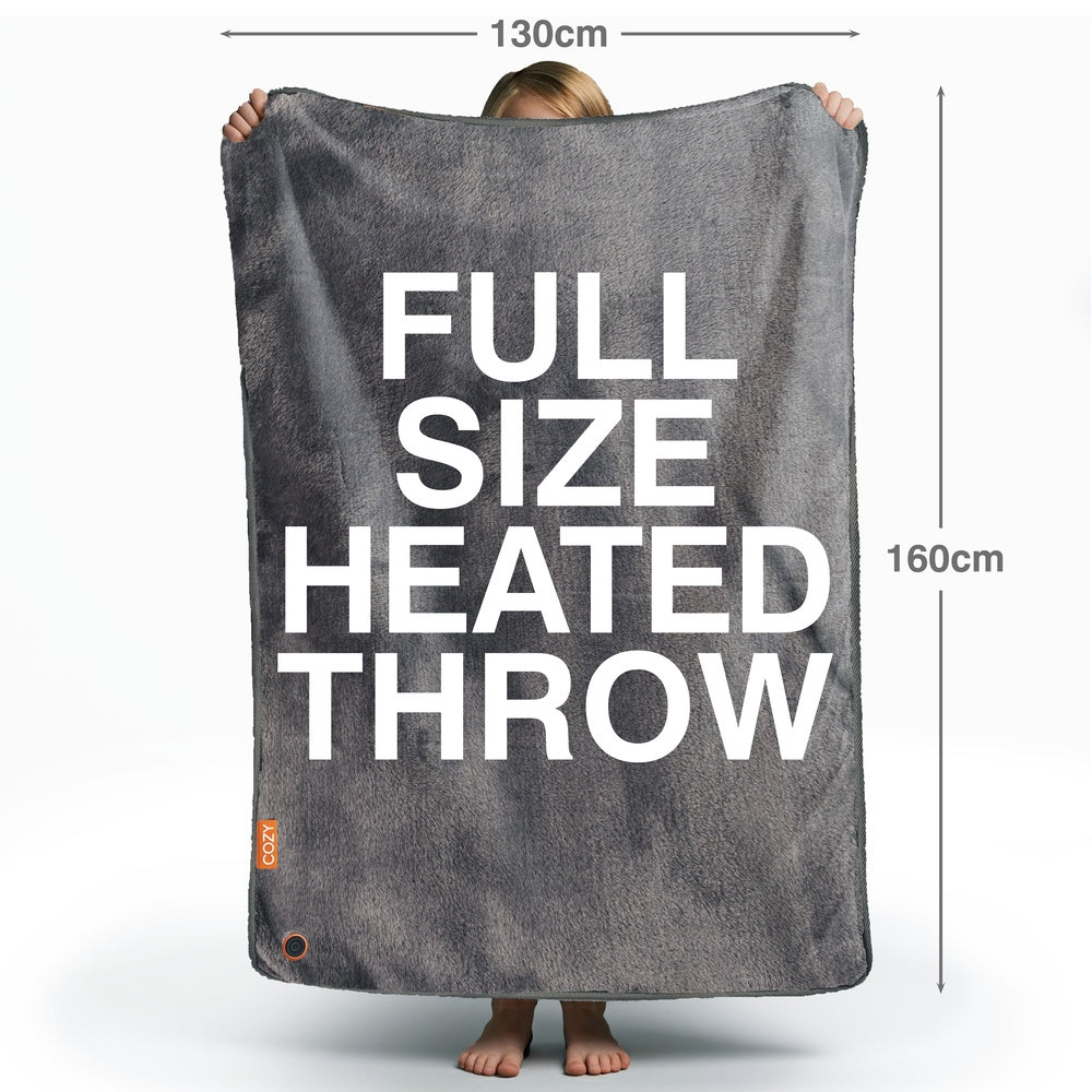 A person holds up a large Cozy Cushion EU Cozy Wireless Rechargeable Snuggle Blanket Heated Throw (130 x 160 cm), with bold white text reading FULL SIZE HEATED THROW. Only the person's feet and part of their head are visible.