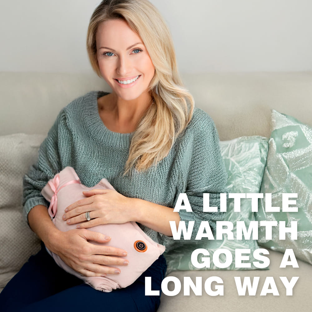 A woman in a green sweater and navy pants sits smiling on a couch, holding the UNA Rechargeable Hot Water Bottle from Cozy Cushion EU to her stomach. Text: A LITTLE WARMTH GOES A LONG WAY.