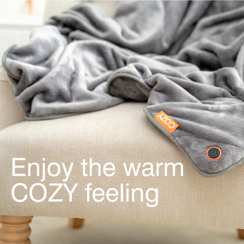 A gray Cozy Cushion EU Wireless Rechargeable Snuggle Blanket Heated Throw with an orange COZY tag is draped over a beige armchair. Large white text reads: Enjoy the warm COZY feeling.